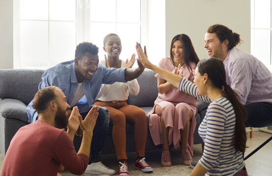 Group Of Happy Young Multiethnic People Sitting In Living Room And Having Fun Together. Two Cheerful Excited Multicultural Multiracial Millennial Friends Giving Each Other A High Five And Laughing