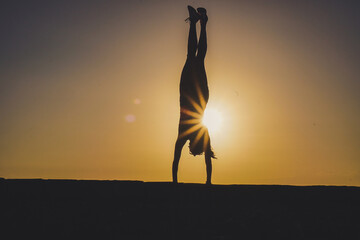 silhouette of a sportive athletic woman doing handstand on beach boardwalk during sunset 
