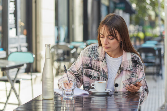 Young woman browsing on mobile phone and making notes in cafe