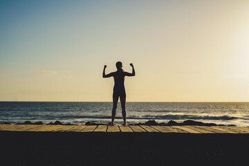 Fototapeta premium silhouette of a successful, strong and sportive female doing champion pose with fists on beach boardwalk during sunset or sunrise 