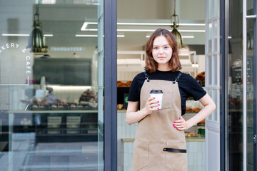 Cute confident baker girl posing with a plastic coffee cup in front of her shop