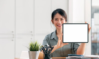 Portrait of young smiley woman showing blank screen tablet while sitting at her office desk.