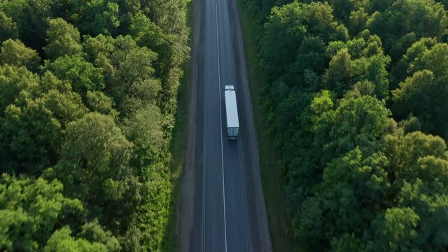 A white semi-truck with an awning trailer crosses the highway located next to the forest. Shooting from the air in the evening. Business in the field of transportation of heavy loads. High quality.