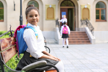 Cute African-American schoolgirl sitting on bench outdoors