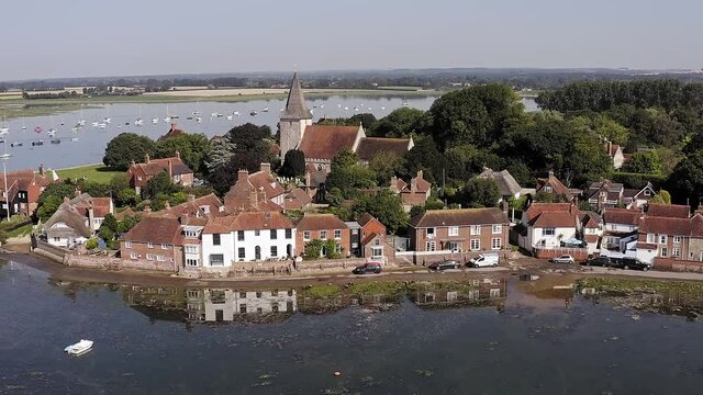 Aerial footage of the historic village of Bosham on an estuary in West Sussex with the Holy Trinity Church in the centre of the village and reflections of the cottages in the water.