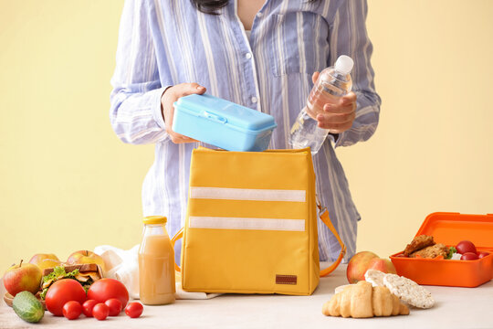 Woman packing meal into lunch box bag on color background
