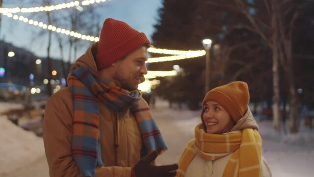 Happy young couple walking on street decorated with garlands, chatting and embracing while having romantic outdoor date in winter