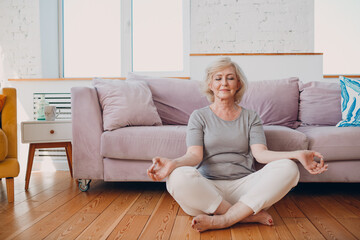 Senior adult smiling woman practicing yoga at home living room. Elderly relaxed female sitting in lotus pose and meditating at home - obrazy, fototapety, plakaty