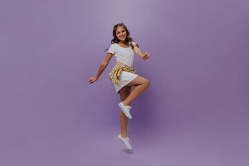 Fototapeta na wymiar Tanned teenager jumping on isolated. Curly happy young girl in white dress and checkered shirt moves on purple background.