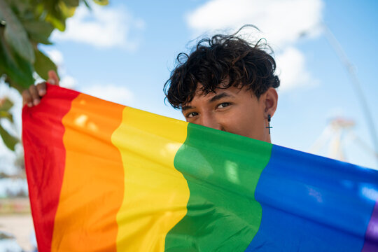 Portrait of young Hispanic gay boy looking at camera, holding LGBT flag - Focus on face landscape imagin