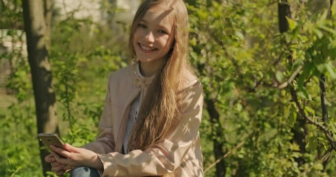 Portrait Smiling preteen girl using mobile phone sitting on the fallen tree in the park and smiling at camera. Girl has long blond hair