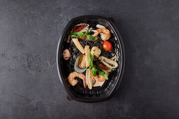 Black spaghetti Nero with seafood, tomatoes and basil in black plastic container on dark background. Designer food concept for take away or delivery