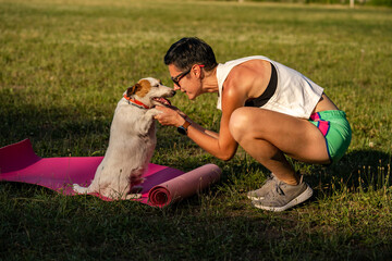 Young sporty woman with short black hair training outdoors on yoga mat on green grass in a park, activity with dog, jack russell terrier walk