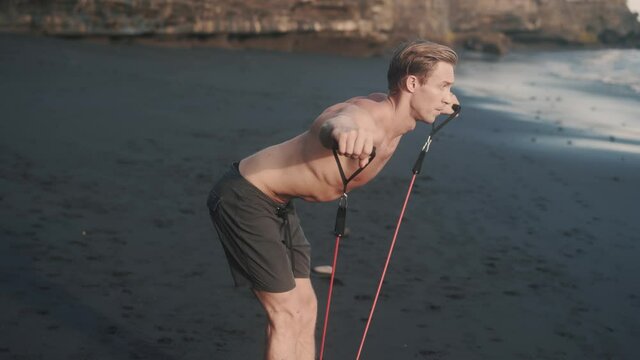 Young fitness Caucasian man does exercise with elastic band at the seashore. Perfect body young guy trains his strong arms and torso with resistance band. Concept of beauty. High quality 4k footage