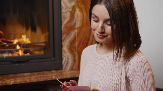 A young woman holds a beautiful bouquet of roses, sitting on a near a burning fireplace. Reads a note with wishes.