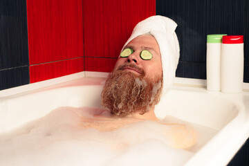 Cute bearded man taking bath with head wrapped in towel and cucumber slices on his eyes. Funny hipster relaxing in foamy bathtub and enjoying life. SPA at home.
