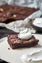 Brownie with ginger, dark chocolate coloured, on a white parchment lying on the gray table. A piece of brownie lies on a plate in the foreground. A bowl of cream stands near. Close up. 45 angle view