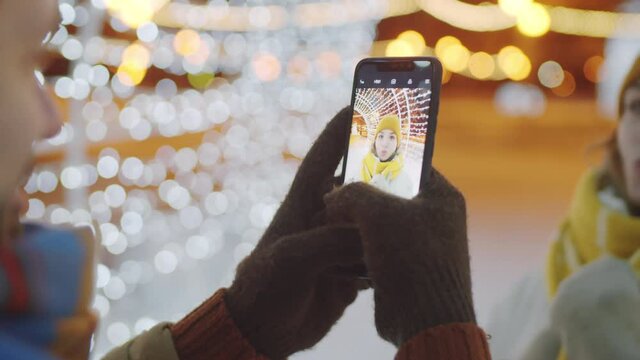 Close up selective focus shot of man taking picture of his cheerful girlfriend in tunnel of Christmas lights outdoors