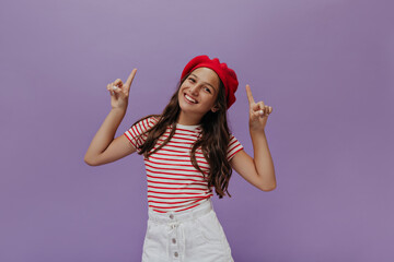 Obraz na płótnie Canvas Happy brunette girl in striped t-shirt points up on isolated purple background. Teenager in red beret sincerely smiles.