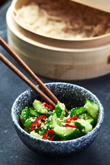 Smashed cucumbers with sesame and chili peppers in a grey bowl on a table with chopsticks. 45 angle view