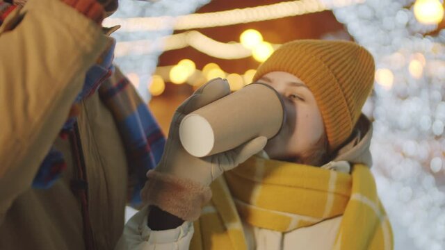 Young loving couple drinking coffee from to go cups, kissing and embracing while having romantic date outdoors on street decorated with Christmas lights