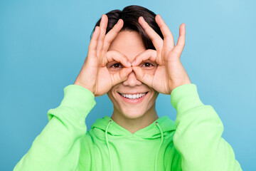 Photo of funny foolish guy show okey sign cover eyes look camera wear green hoodie isolated blue color background