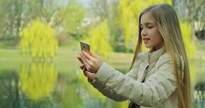 Smiling preteen girl making selfie using mobile phone near pond in the park and smiling at camera. Girl has long blond hair