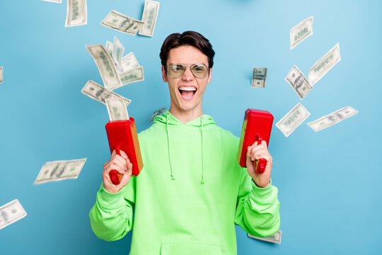 Photo of funky cool young guy dressed green hoodie shooting money gun smiling isolated blue color background