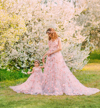 Happy beautiful family mom and daughter, spring blooming garden. Long luxury evening fantasy dresses powder pink color. Queen gently holds little princess hand. Hairstyle decorated wreath flowers