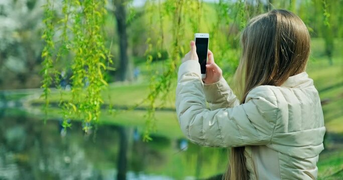 Smiling preteen girl shooting photo with mobile phone when sitting on the grass near pond in the park. Girl has long blond hair looking at camera