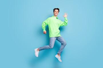 Fototapeta na wymiar Photo of sweet attractive young gentleman wear green sweatshirt smiling jumping high waving arm isolated blue color background