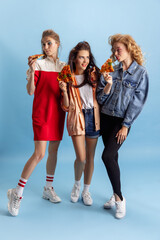 Three young attractive women in retro 90s fashion style, outfits eating pizza isolated over blue...