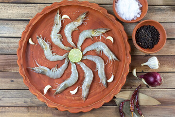 Fresh raw shrimps in a earthen plate along with onion, garlic and spices on wooden background.