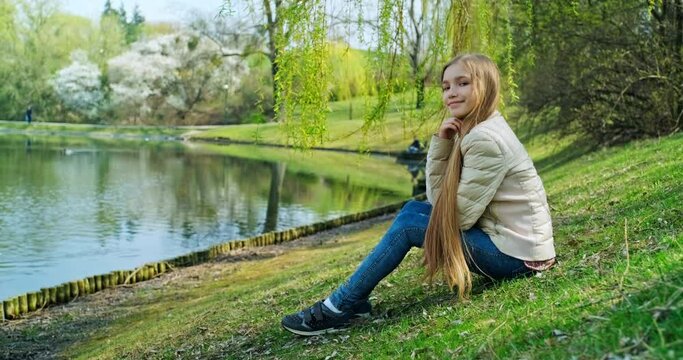 Cheerful preteen girl sitting on the grass near pond in the park. Girl has long blond hair smiling at camera