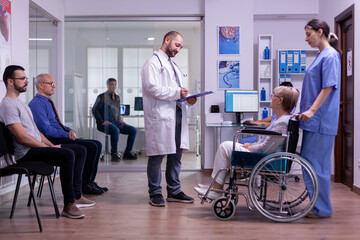 Specialist doctor checking diagnosis of disabled senior woman in wheelchair reading on clipboard. Professional medical staff in hospital hallway with invalid patient, man in medical examination room