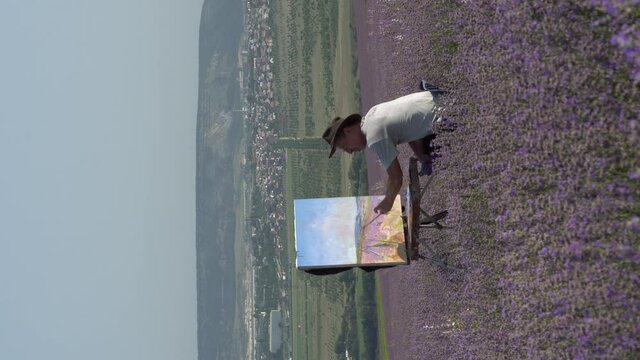 Adult artist man in a hat paints a picture on canvas in a lavender field. Open air in nature, rural landscape. Hobbies recreation and art therapy, oil painting. Vertical video