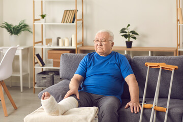 Unhappy disappointed senior mature man sitting on sofa with broken leg in cast and crutches nearby...