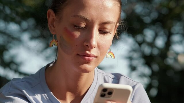 A close-up view of a beautiful woman with rainbow make-up is typing message using her phone during pride gay parade outside
