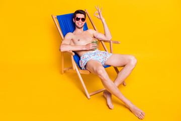 Full length body size view of attractive cheery guy sitting in chair showing ok-sign drinking juice isolated over bright yellow color background