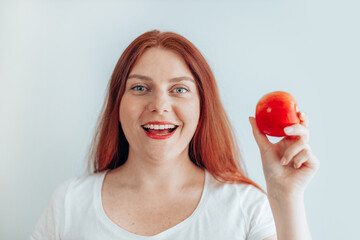 Portrait of a cheerful young woman in a white t-shirt eating red apple on gray wall background. Healthy nutrition diet. Apple vitamin snack.