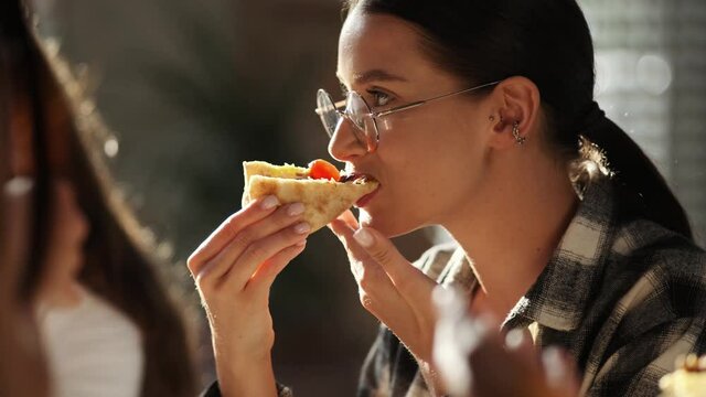 A close-up view of a Caucasian business woman wearing glasses is eating pizza with her colleagues in the office