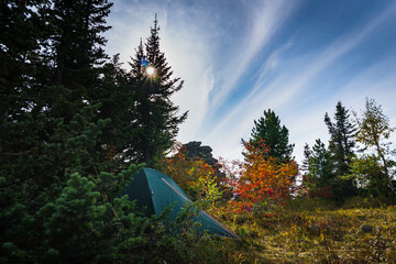 A tourist tent standing on the top of a mountain in the autumn forest against the backdrop of a blue cloudy sky and the setting sun