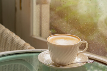 Cup of latte coffee with latte art close to the windows with rain drop