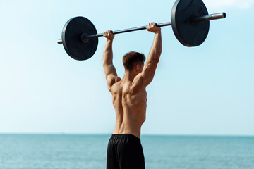 Muscular man training on the beach by the sea, doing exercises with a barbell for biceps, strong male press with a naked torso, sports concept - obrazy, fototapety, plakaty