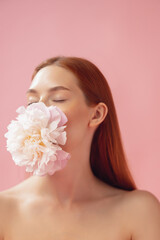 Obraz na płótnie Canvas Close-up portrait of beautiful redheaded woman with flower isolated over pink studio back ground. Nude color, diet, cosmetics, natural beauty and aesthetic cosmetology concept.