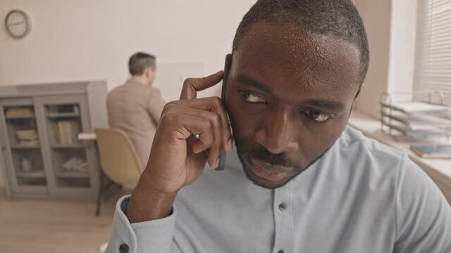 High angle close-up of young anxious African manager talking on mobile phone in office, sweating