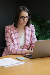 European confident business woman in a pink jacket and with a laptop in her office working, modern young business woman director working in her office and talking on the phone with colleagues, online