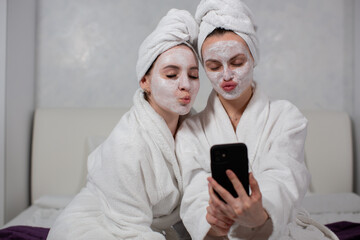 Two funny girl girlfriends in bathrobes, face masks and towels on their heads take selfies on the phone. High quality photo