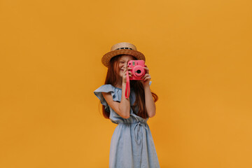 Long-haired redhead girl in boater and denim blue dress holds pink camera and takes photo on orange isolated background.