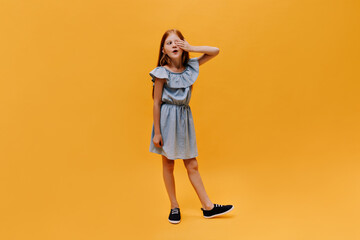 Full-length portrait of redhead charming young girl in denim dress and black sneakers closing one eye and posing on orange background.